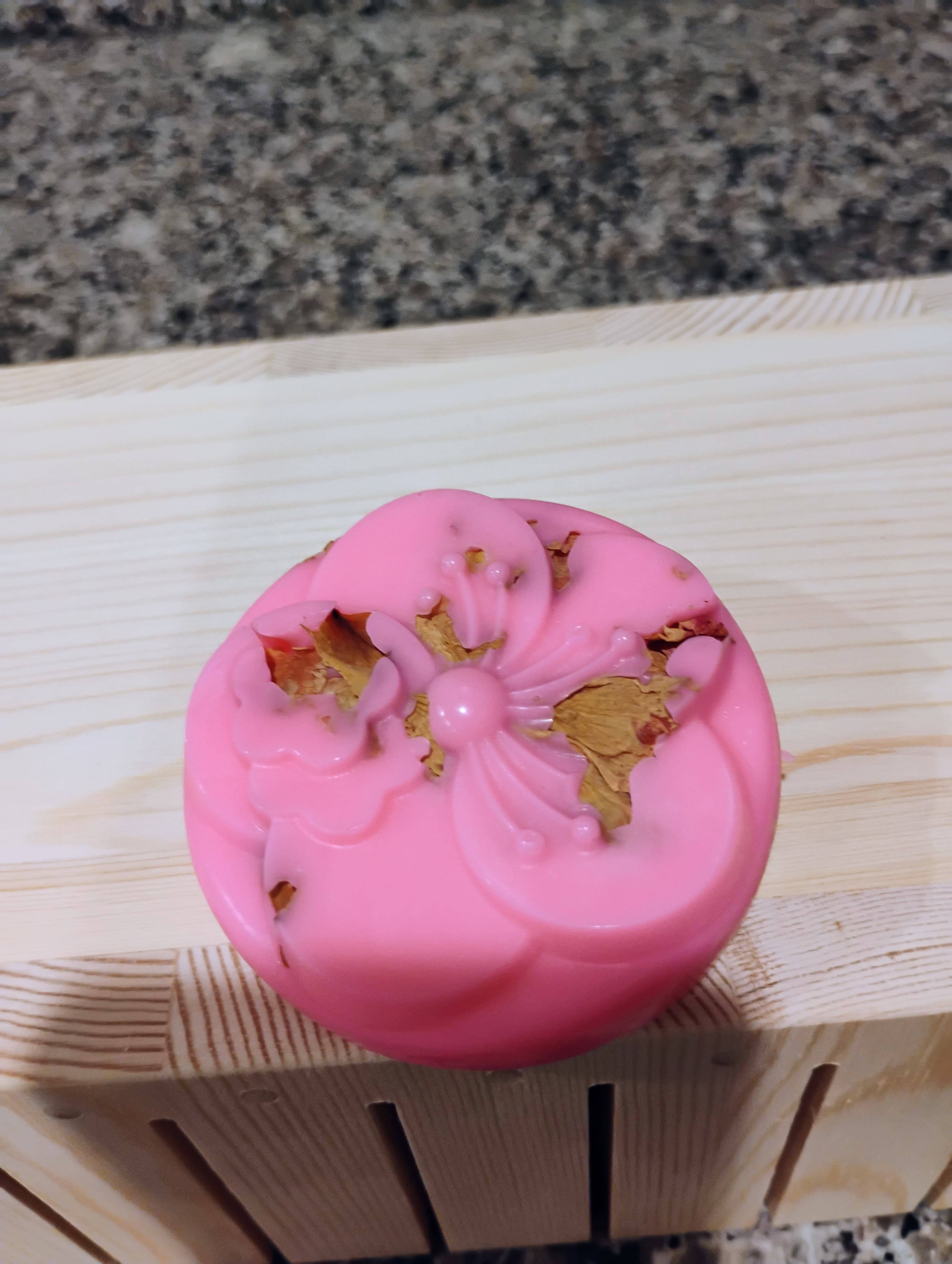 Round pink soap with a molded cherry blossom design and embedded dried petals on wood.